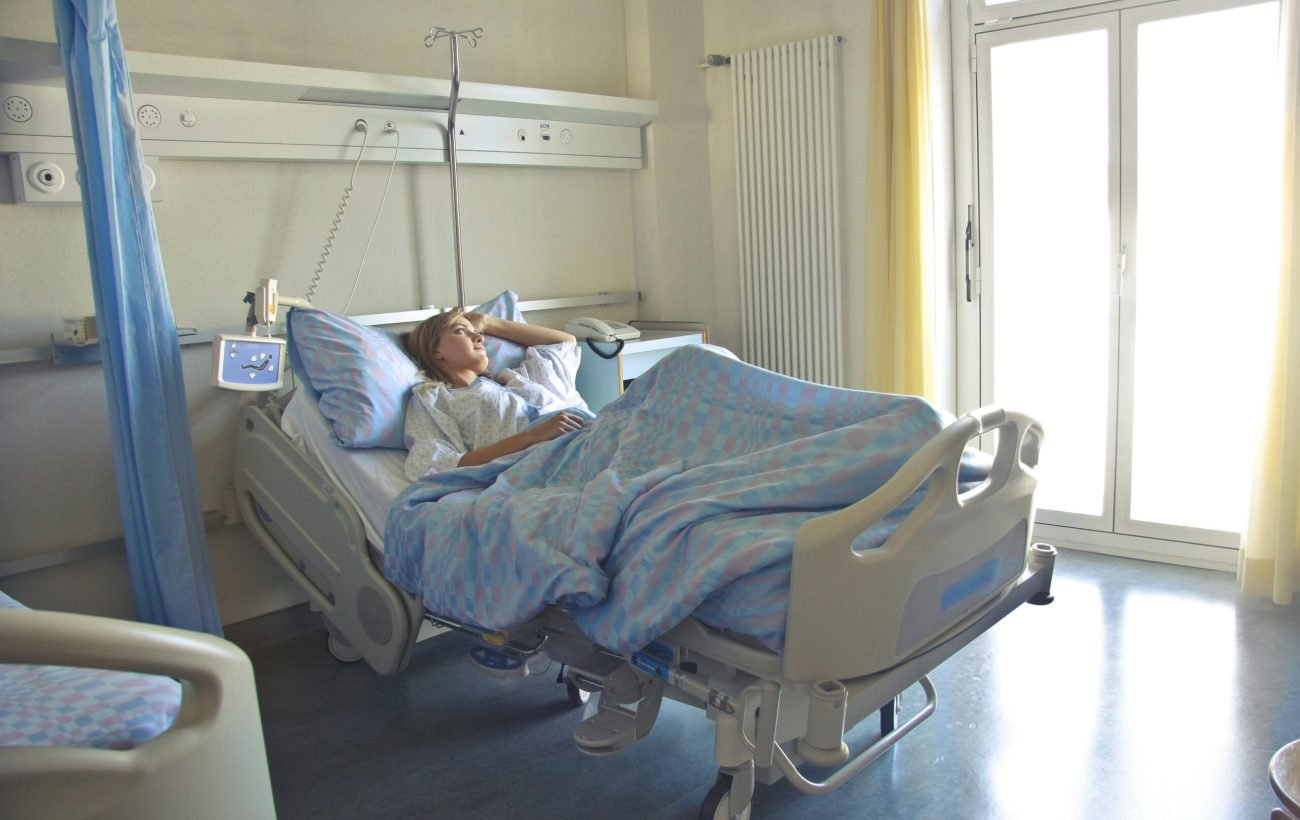 A woman lying in a hospital bed in a bright, softly lit room, appearing to rest or recover.